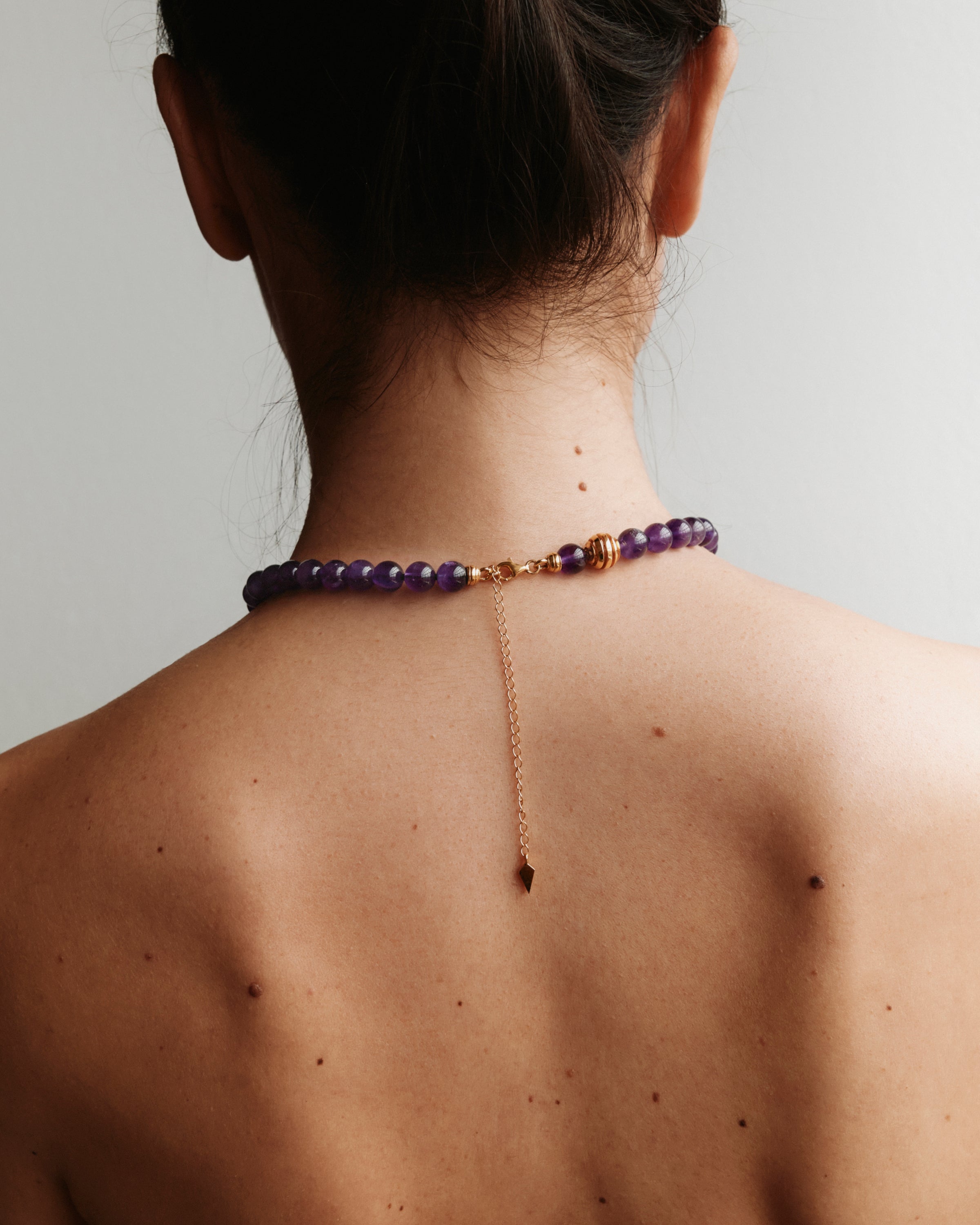 ETERNA VÉLA Amethyst necklace with polished gold spiral bead, 8 cm extension chain and clasp, arrowhead-shaped fantasy signature, and 8 mm faceted amethyst bead strand.