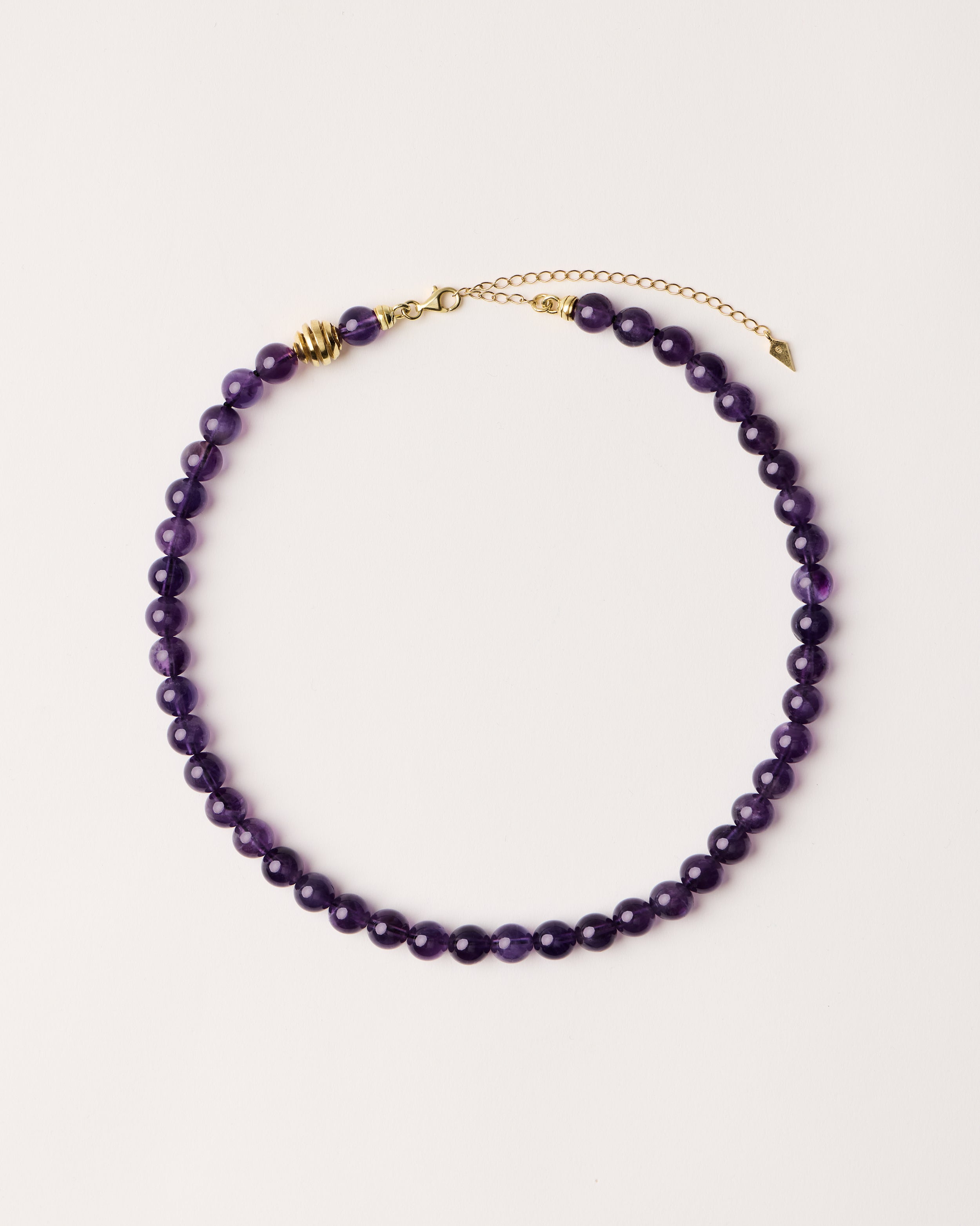 ETERNA VÉLA Amethyst necklace with polished gold spiral bead, 8 cm extension chain and clasp, arrowhead-shaped fantasy signature, and 8 mm faceted amethyst bead strand.